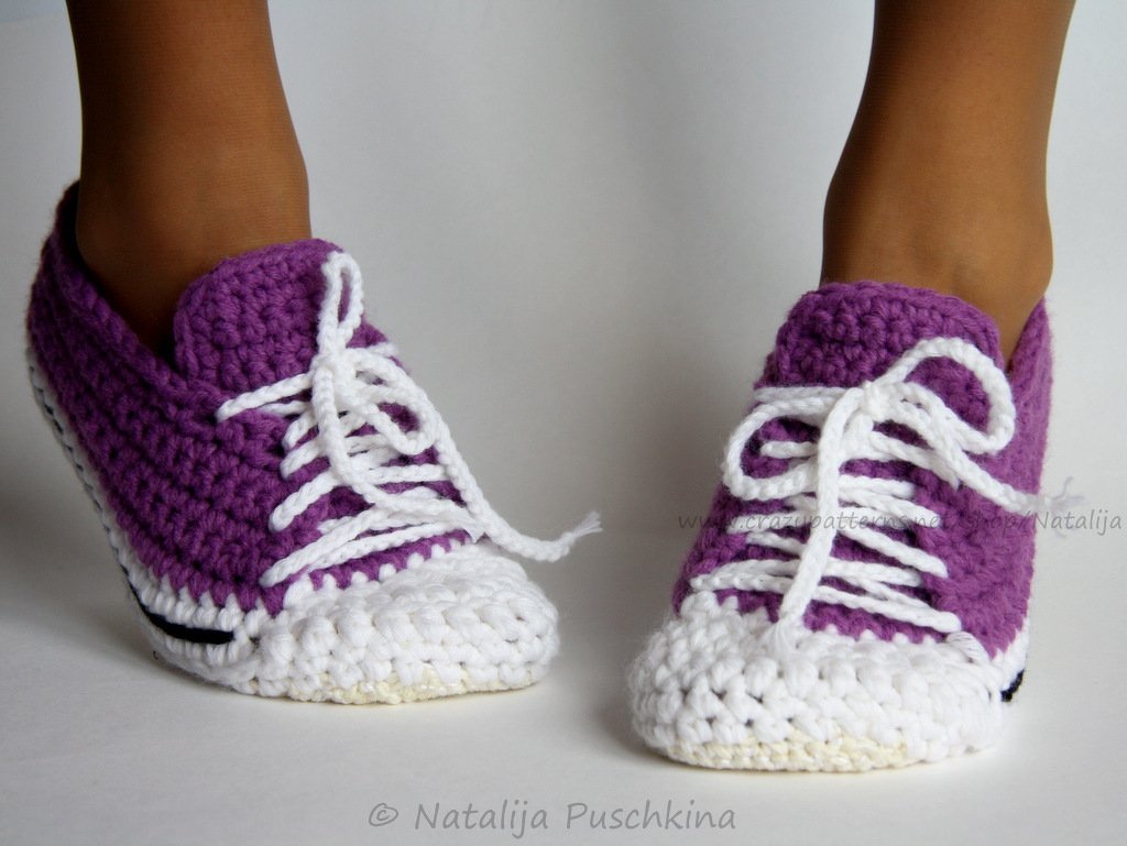 Lila-weiße, gehäkelte Hausschuhe in Sneaker-Form mit weißen Schnürsenkeln an Füßen