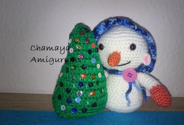 Gehäkelter weißer Schneemann mit blauer Kapuze und rosa Knopf neben grünem gehäkeltem Weihnachtsbaum mit bunten Pailletten
