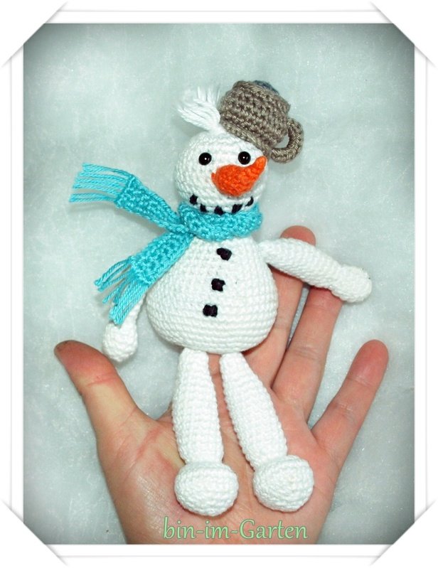 Hand hält kleinen gehäkelten Schneemann mit türkisfarbenem Schal, oranger Nase und brauner Mütze