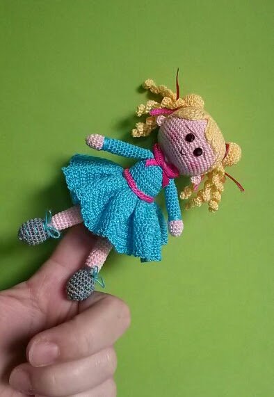 Gehäkelte Amigurumi-Fingerpuppe mit blauem Rüschenkleid und blonden Locken auf einem Finger.