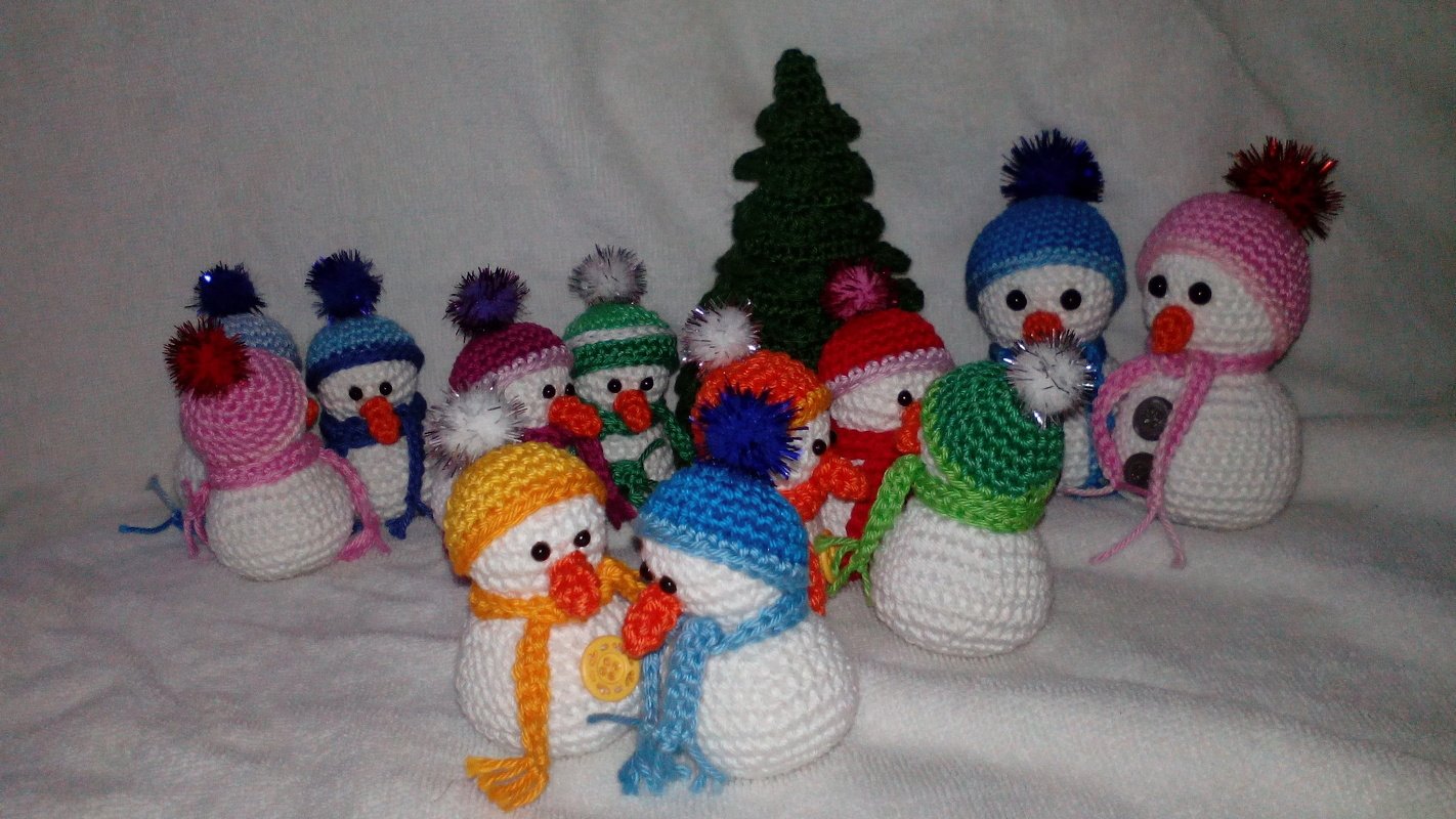 Mehrere gehäkelte Amigurumi-Schneemänner mit bunten Mützen und Schals vor einem kleinen gehäkelten Tannenbaum