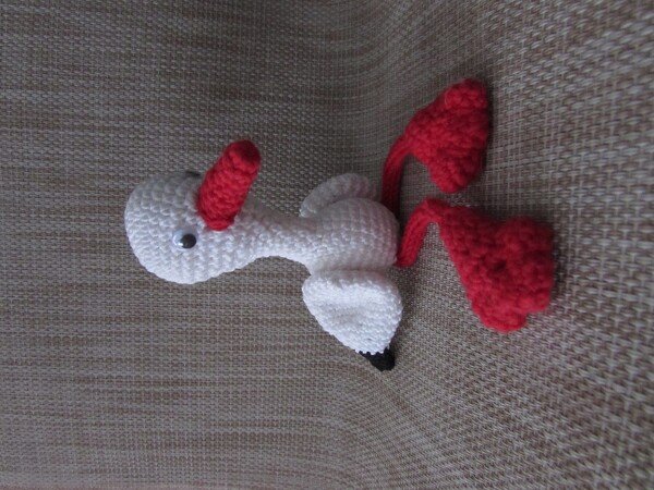 Gehäkelter Amigurumi-Vogel seitlich liegend auf beigem Stoff, mit roten Beinen und rotem Schnabel