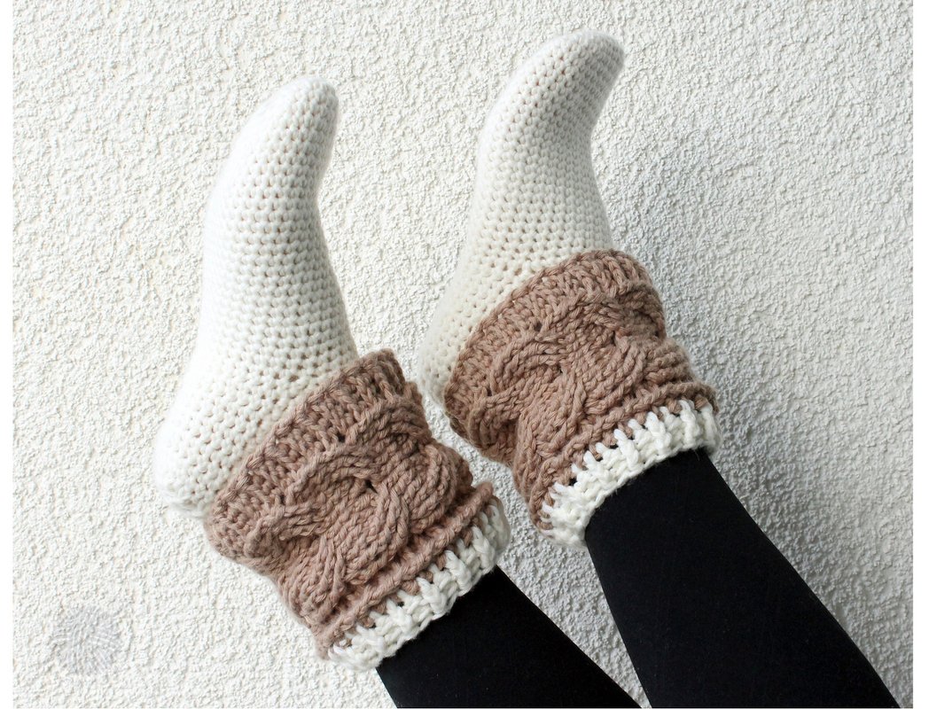 Beine in schwarzen Leggings mit cremeweißen Häkelsocken und beigebraunen, zopfartigen Stulpen vor heller, strukturierter Wand.