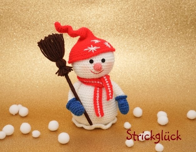 Crocheted snowman amigurumi with red hat, striped scarf, blue mittens, holding a broom on gold background with white pom-pom snowballs