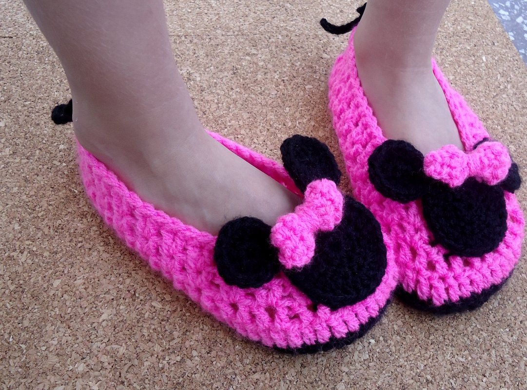 Child wearing bright pink crocheted slip-on slippers with black mouse-shaped appliqués and small pink bows