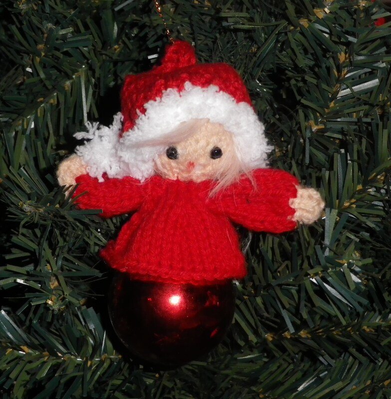 Gestrickter roter Amigurumi-Engel mit weißem Mützensaum auf roter Kugel vor Tannengrün