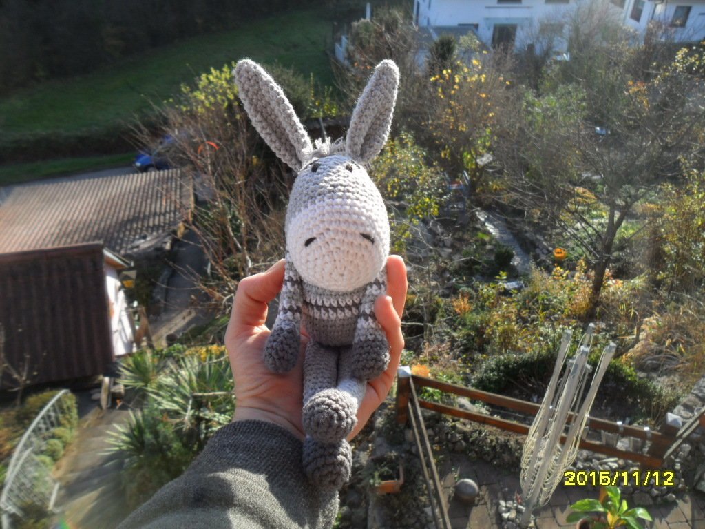 Handgehaltener, gehäkelter Amigurumi-Esel in Grau und Weiß vor unscharfem Landschaftshintergrund.