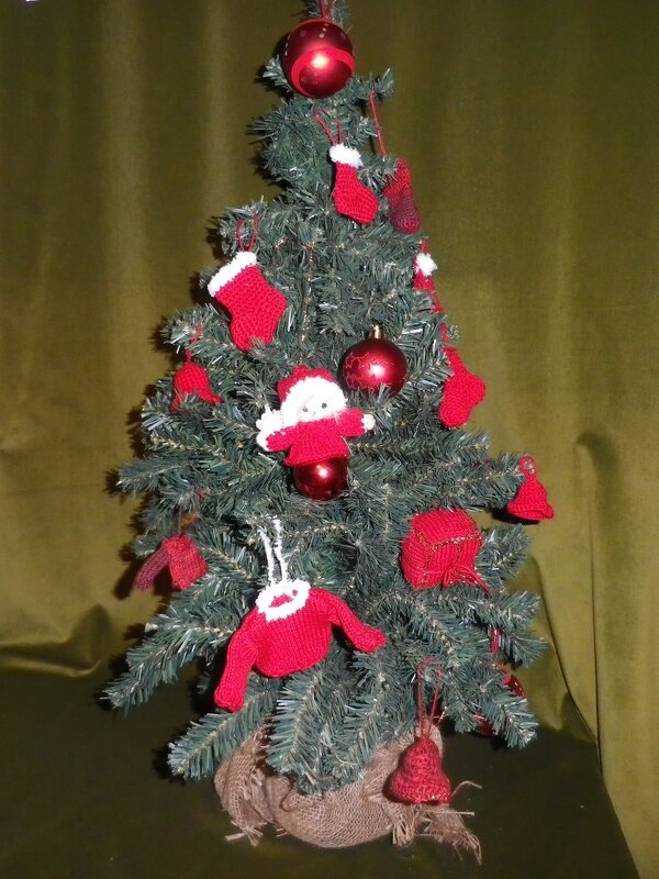 Künstlicher Weihnachtsbaum mit selbstgemachtem, roten Häkelschmuck wie kleinen Pullovern, Socken und einer Weihnachtsmann‑Figur.