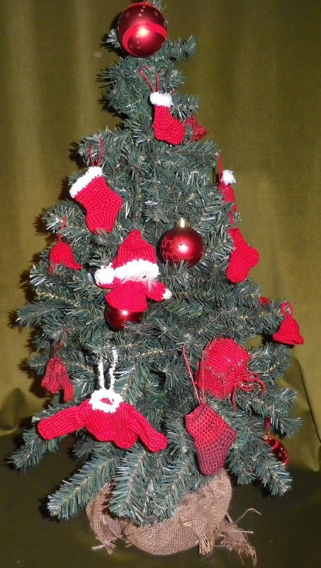 Kleiner künstlicher Tannenbaum mit roten, gehäkelten Mini-Stiefeln, einer Nikolausfigur, einem Pullover und roten Kugeln, der Stamm ist in Jute umwickelt.
