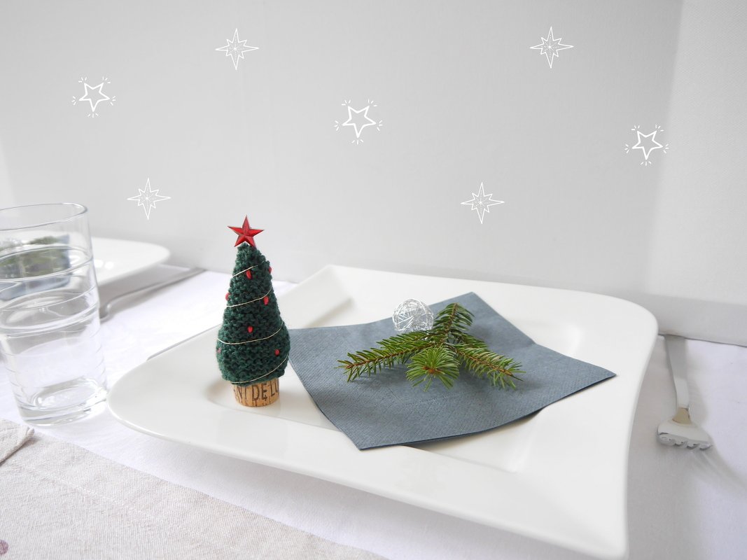 kleiner gestrickter Tannenbaum auf Korksockel auf weißem Teller neben Serviette und Tannenzweig
