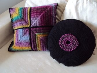 Zwei gehäkelte Kissen auf einem hellen Sofa: quadratisches buntes Quadratmuster und rundes schwarzes Schallplatten-Design.