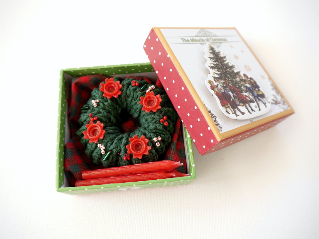 Gehäkelter dunkelgrüner Adventskranz mit roten Stern-Kerzenhaltern und Stabkerzen in offener Geschenkbox auf kariertem Stoff.