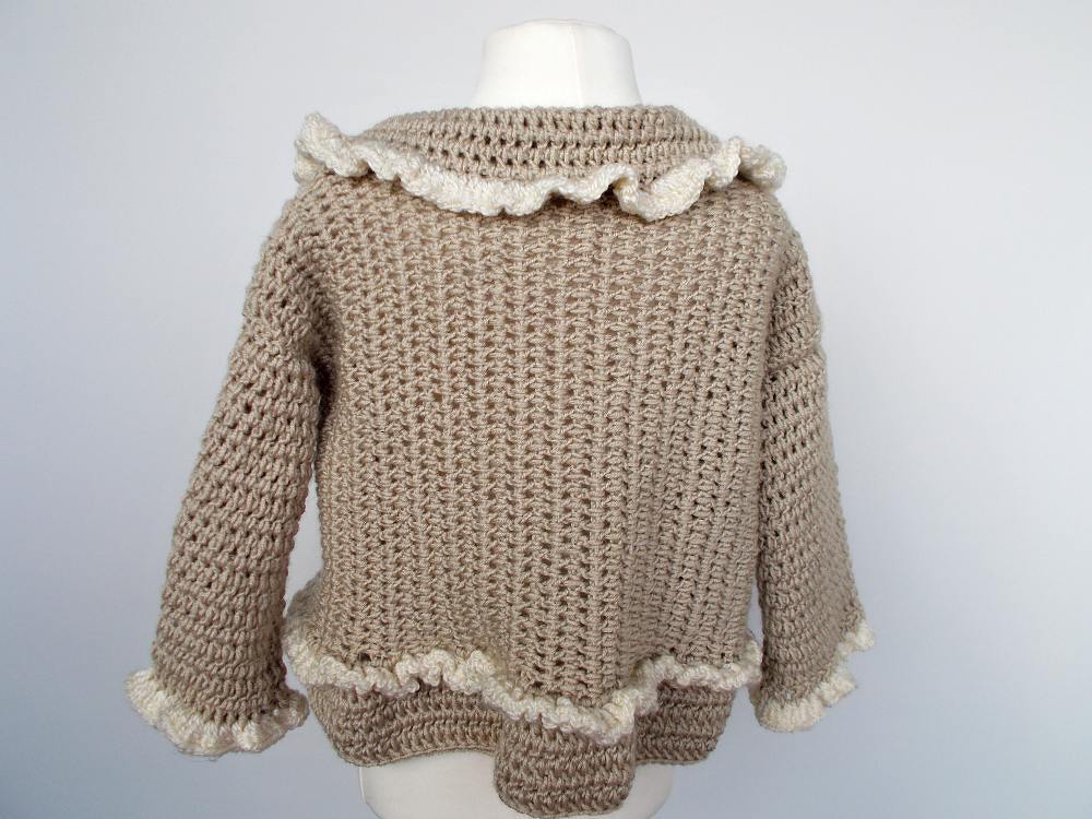 Rückansicht einer beigen gehäkelten Babyjacke mit cremefarbenen Rüschen an Kragen, Ärmeln und Saum auf einer Schaufensterpuppe.