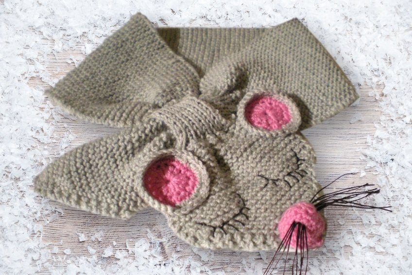 Grey knitted mouse-shaped scarf with pink inner ears and a pink nose, black whiskers and embroidered closed eyes on a wooden surface with white flakes.
