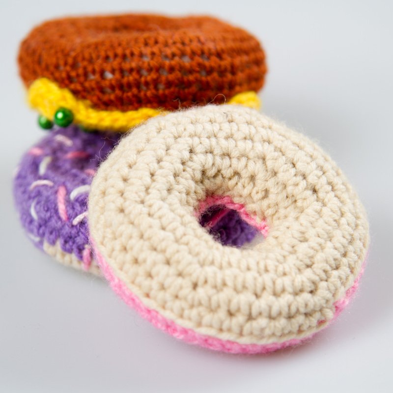 Drei gehäkelte Amigurumi-Donuts in Creme, Lila und Braun mit unterschiedlichen Verzierungen