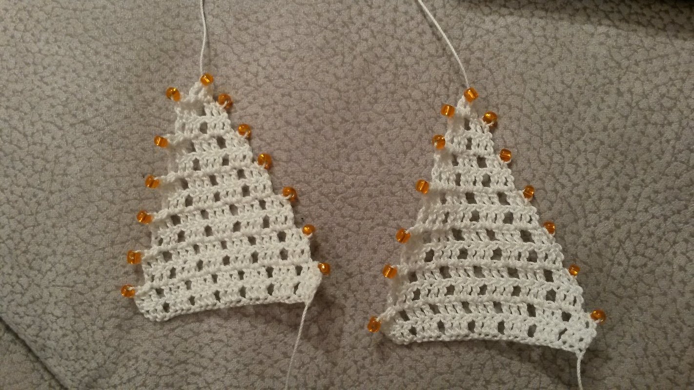 Zwei weiße, gehäkelte Weihnachtsbaumformen mit orangefarbenen Perlen an den Rändern auf grauem Untergrund