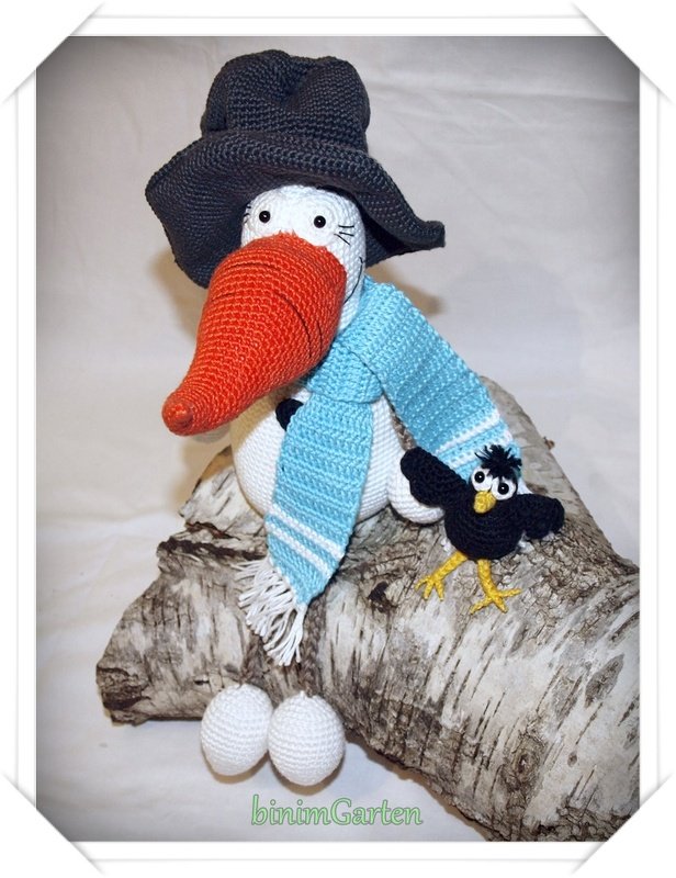 Gehäkelter weißer Amigurumi-Vogel mit großem orangefarbenem Schnabel, schwarzem Hut, hellblauem Schal, kleiner schwarzer Krähe und zwei gehäkelten Eiern auf einem Birkenstamm