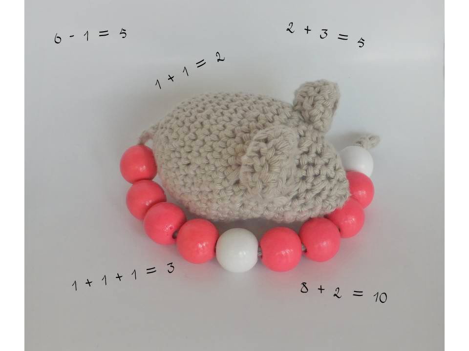 Gehäkelte Maus mit korallenroten und weißen Rechenperlen und einfachen Rechenaufgaben auf hellem Hintergrund.