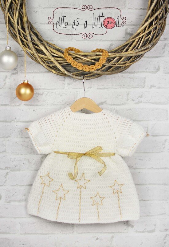 Weißes gehäkeltes Babykleid mit goldenen Sternen und goldfarbenem Satinband an einem Holzhänger vor weißer Ziegelwand.