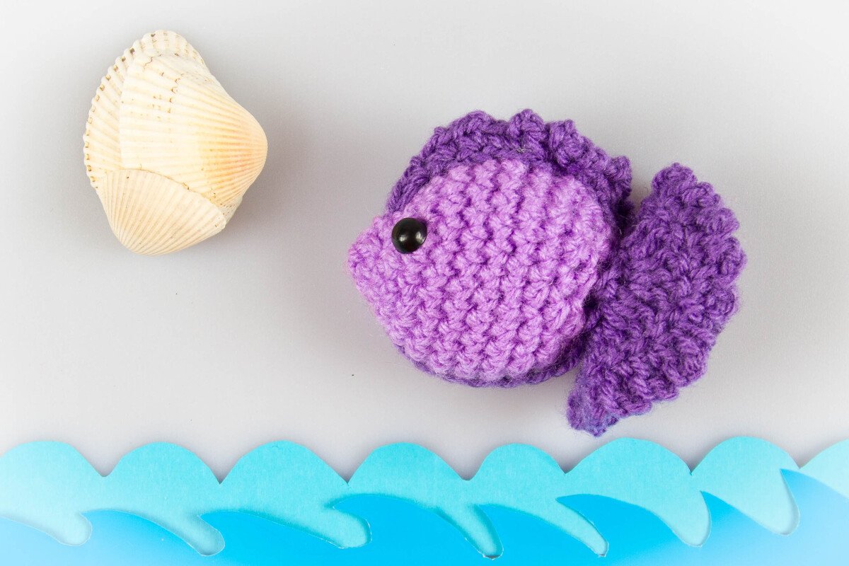 Violetter gehäkelter Amigurumi-Fisch mit schwarzem Auge neben Muschel und blauen Papierwellen