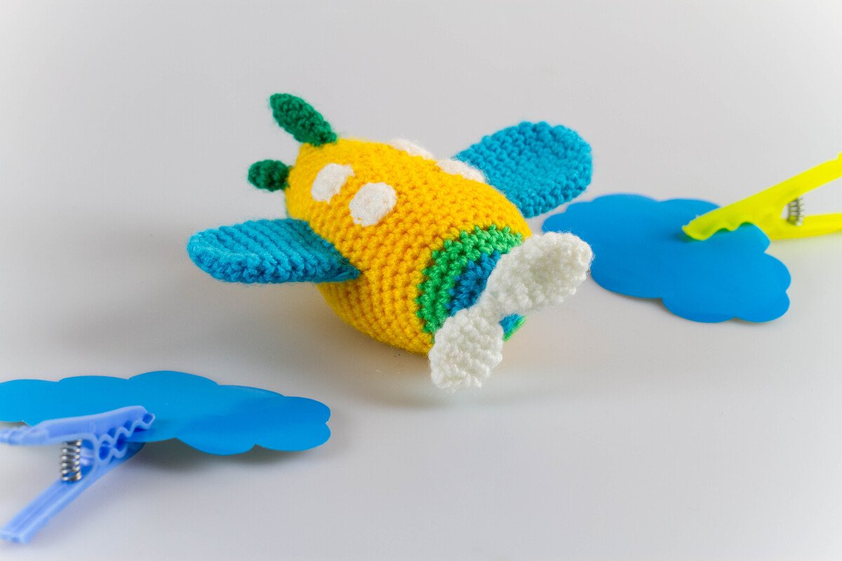Gehäkeltes Amigurumi-Flugzeug in Gelb mit blauen Flügeln, weißem Propeller und grünen Akzenten auf hellem Hintergrund.