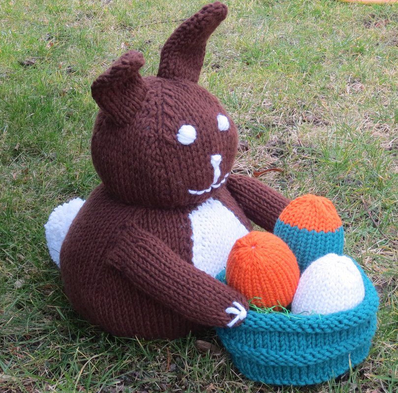 Runder brauner Amigurumi-Hase mit weißer Bauchpartie sitzt im Gras und hält einen türkisen Korb mit orange-blauen und weißen gestrickten Eiern