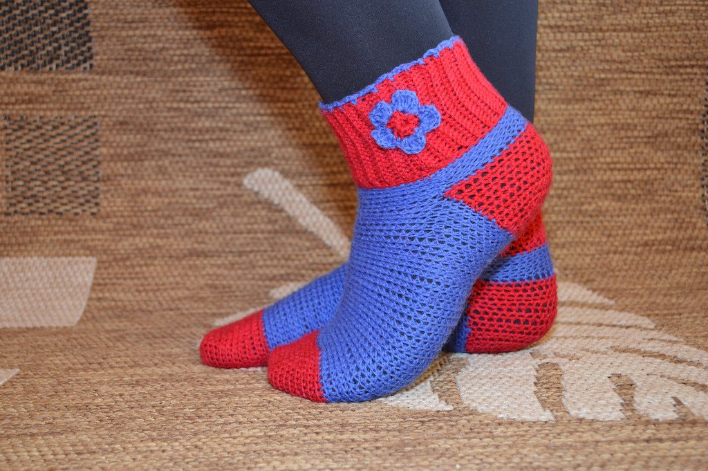 Kurze gehäkelte Socken in Rot und Blau mit blauer Blumenapplikation am Schaft, getragen über dunkle Leggings auf gemustertem Teppich