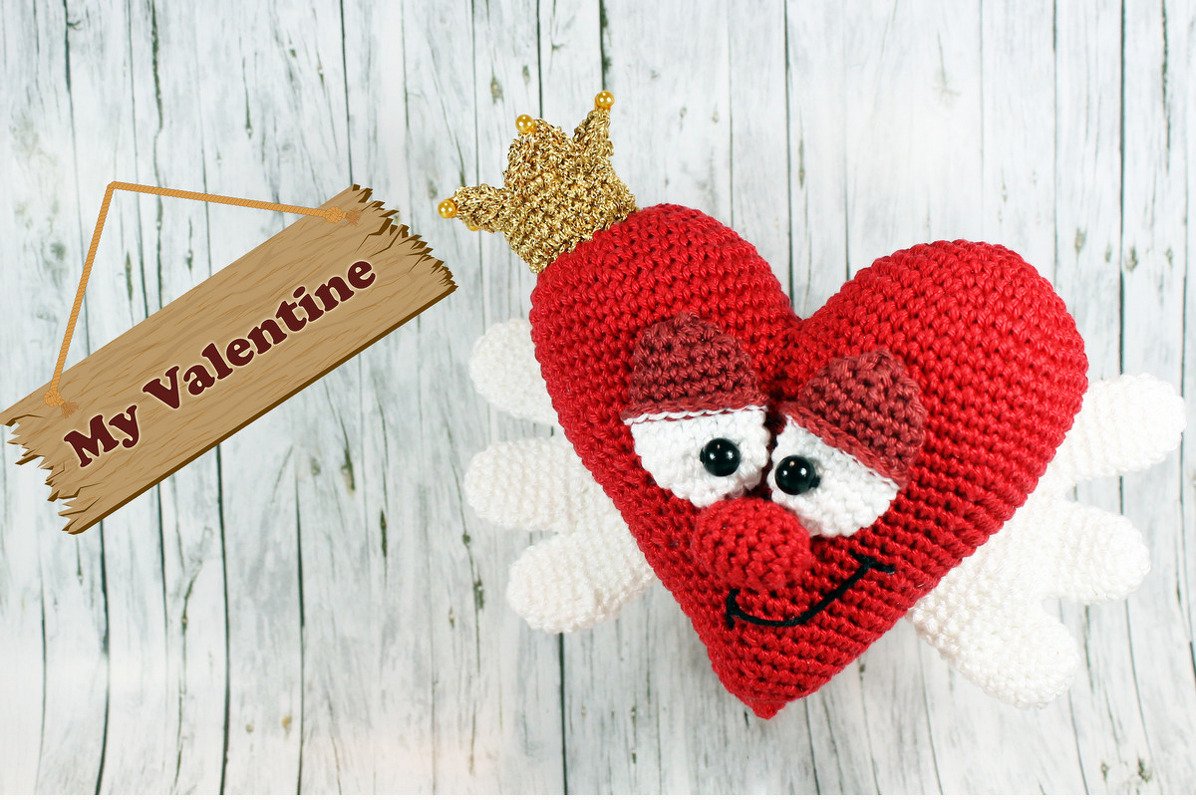 Rotes gehäkeltes Amigurumi-Herz mit weißen Flügeln, schwarzen Knopfaugen und goldener Krone auf hellem Holzuntergrund mit Schild 'My Valentine'