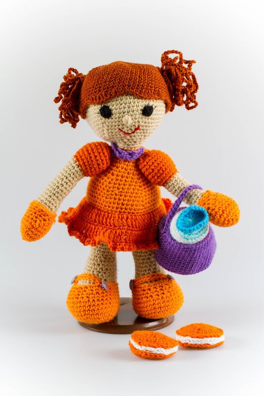 Gehäkelte Amigurumi-Puppe mit rotbraunen Zöpfen, orangefarbenem Kleid, lila Tasche und zwei gehäkelten Keksen