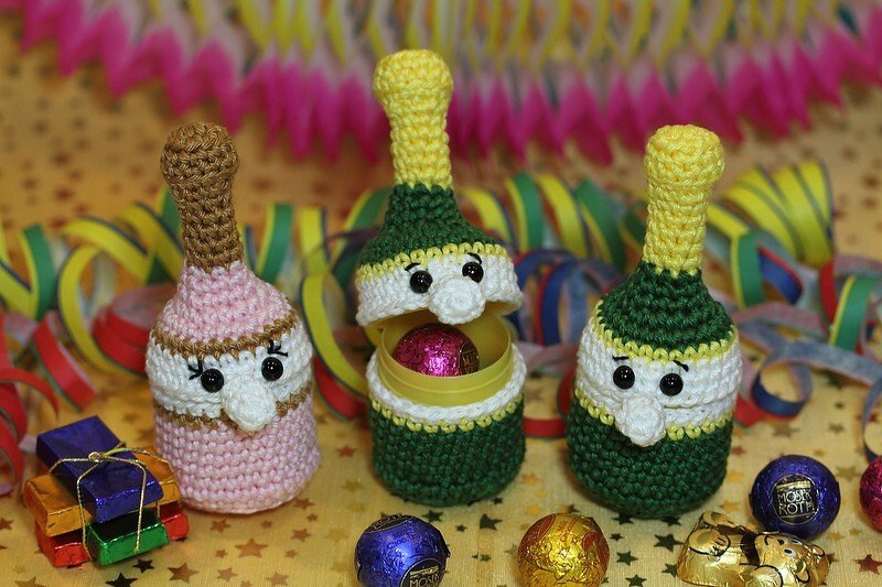 Drei gehäkelte Sektflaschen-Amigurumi vor bunter Partydekoration; eine geöffnet mit Süßigkeit im Inneren.