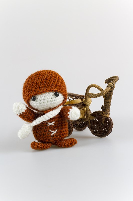 Gehäkelte Amigurumi-Puppe in Braun und Weiß neben einem kleinen Korb-Dreirad