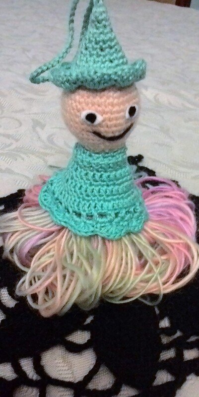 Amigurumi‑Püppchen mit mintgrünem Hut und Rock aus pastellfarbenen Garnfransen auf dunklem Häkeluntergrund