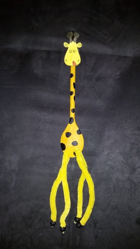 Handgebastelte Giraffe aus gelbem Holzlöffel mit schwarzen Punkten, Pfeifenreinigern als Beine und Pappkopf vor dunklem Hintergrund