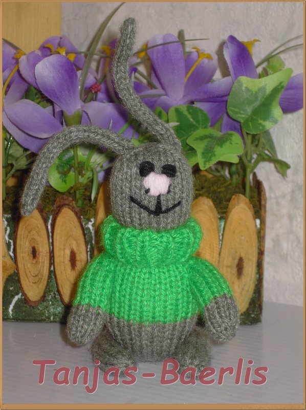 Gestricktes graues Amigurumi-Häschen mit grünem Pullover und rosa Nase vor violetten Blumen