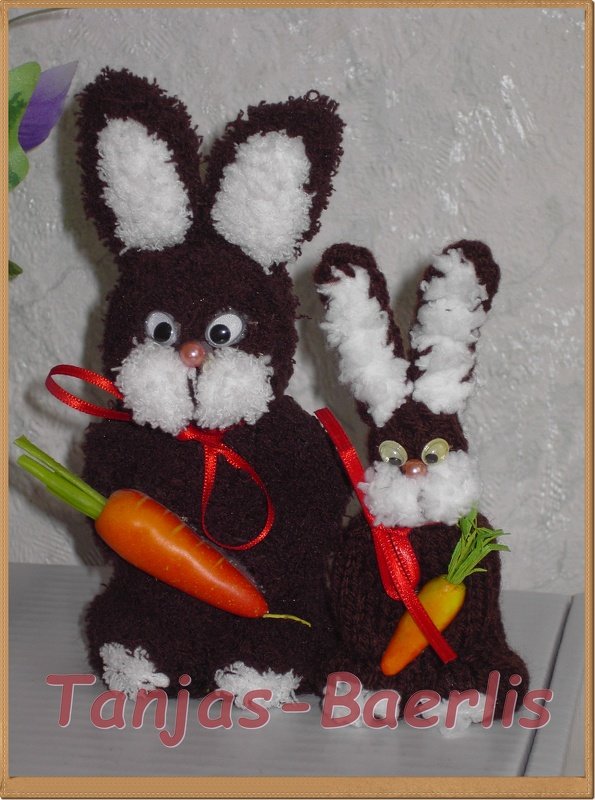 Zwei selbstgemachte, gestrickte Hasenfiguren in Dunkelbraun mit weißen Akzenten, roten Schleifen und kleinen Karotten.