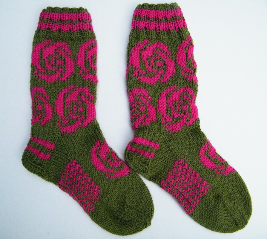 Zwei handgestrickte Socken in Olivgrün mit pinken Rosenmotiven auf weißem Hintergrund