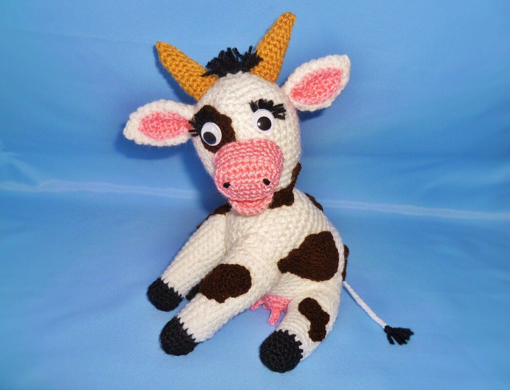 Gehäkelte Amigurumi-Kuh mit braunen Flecken, rosa Schnauze und gelben Hörnern vor blauem Hintergrund