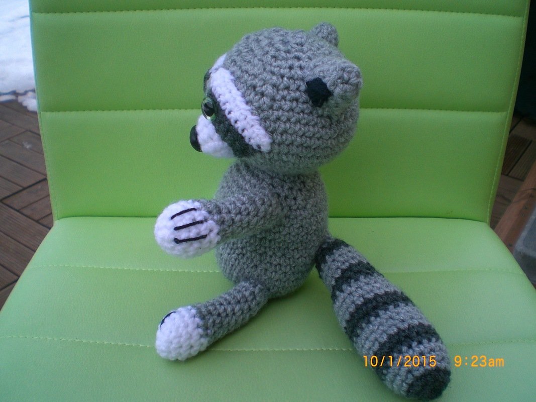 Handgemachter grauer und weißer Amigurumi-Waschbär mit gestreiftem Schwanz auf grünem Stuhl