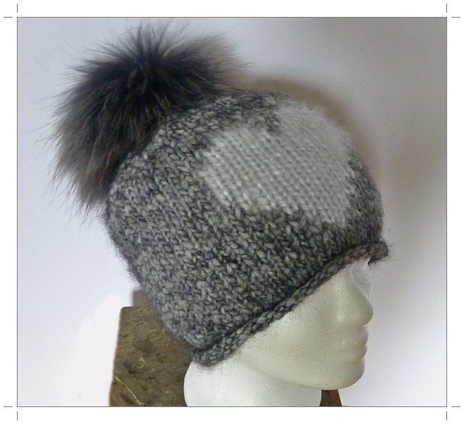 Grau-weiß melierte Strickmütze mit Strukturmuster und großem dunklem Bommel auf Schaufensterpuppe.