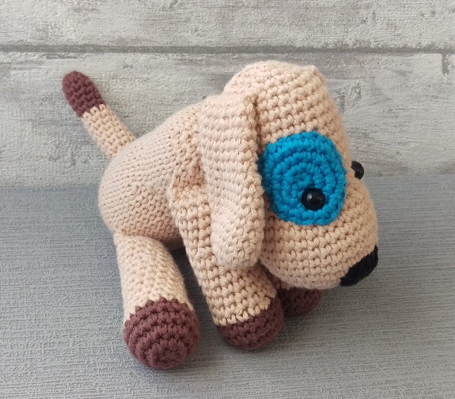 Beiger gehäkelter Amigurumi-Hund mit blauem Augenring, braunen Pfoten und schwarzer Nase