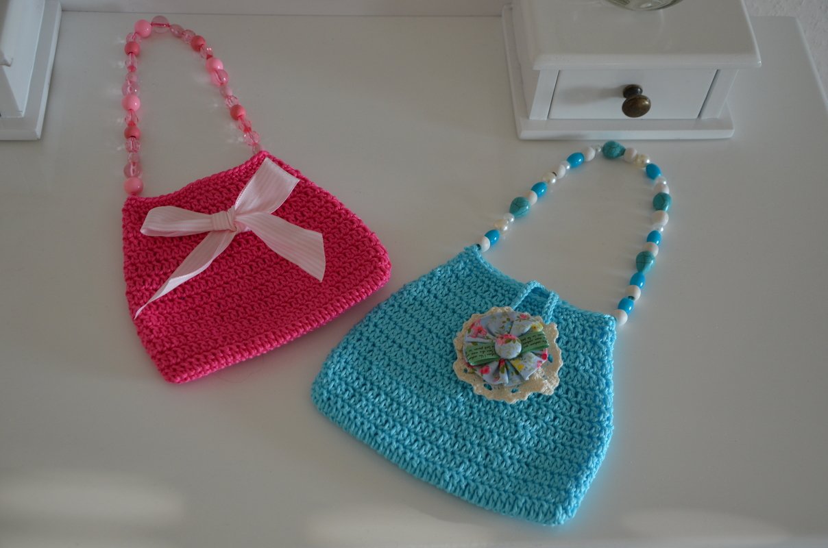 Zwei gehäkelte Henkeltaschen auf weißem Untergrund: pinke Tasche mit Stoffschleife und türkise Tasche mit mehrlagiger Blütenapplikation, beide mit Perlenhenkeln