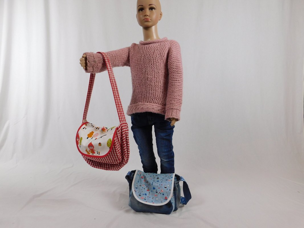 Kinderpuppe mit einer rot-weiß karierten Umhängetasche und einer zweiten blauen Tasche mit Bootsmotiven auf dem Boden.