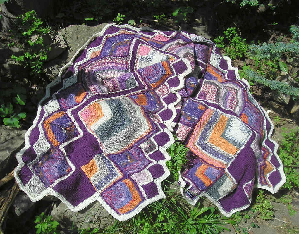 Gestrickter lila Schal mit bunten Quadraten und weißer gezackter Kante auf Felsen und Pflanzen.