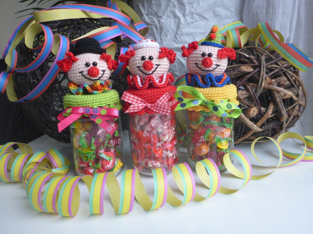 Drei gehäkelte Clown-Köpfe auf Bonbon-Gläsern, umgeben von bunten Luftschlangen.