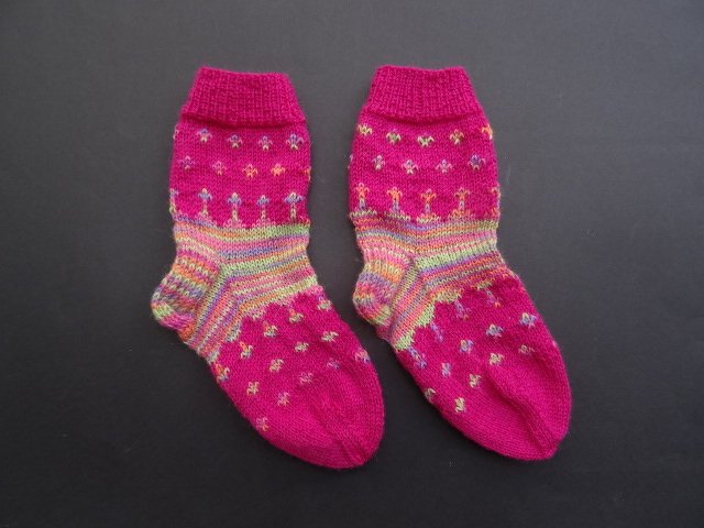 Ein Paar fuchsiafarbene gestrickte Strümpfe mit bunten Streifen und kleinen Mustern auf dunklem Untergrund.