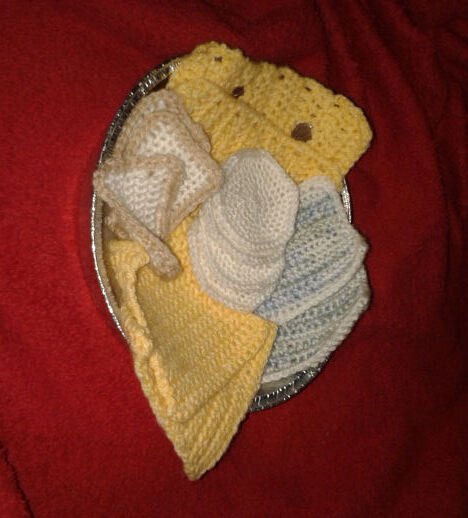 Silberne Schale mit gehäkelten Babyartikeln in Gelb, Beige und Hellblau auf rotem Stoff.
