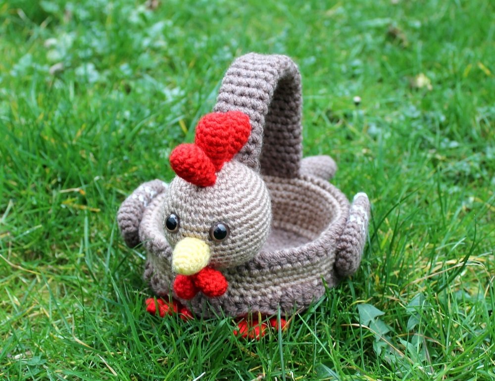 Brown crocheted chicken basket with red comb and wattle, yellow beak and handle, placed on green grass.