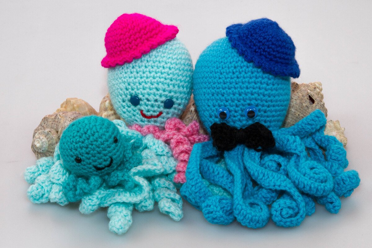 Drei gehäkelte Amigurumi-Oktopusse in Blau- und Türkistönen mit Hüten und Fliege auf Muscheln