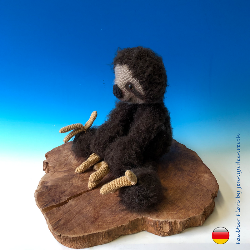 Gehäkeltes Faultier-Amigurumi in dunkelbraun mit beigefarbenem Gesicht und hellen Krallen auf einer Holzscheibe vor blauem Hintergrund.