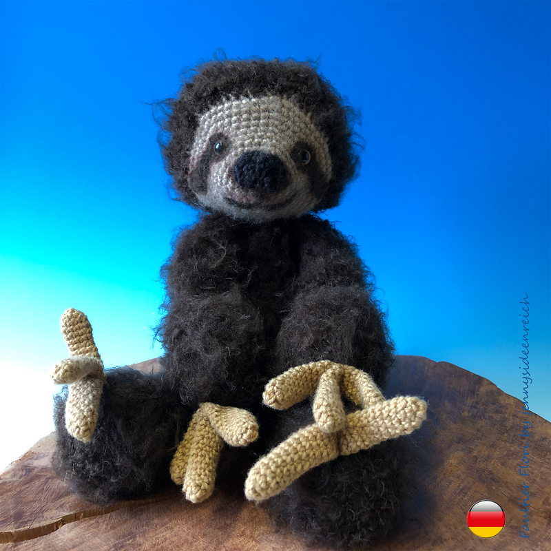 Gehäkeltes Amigurumi‑Faultier mit dunklem, flauschigem Körper und beigefarbenen Krallen auf einer Holzplatte vor blauem Himmel.